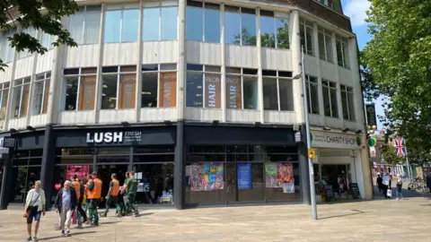 An exterior shot of a post-war building with concrete panels separating the windows of the upper floors. There are a number of retail units on the ground floor, including a Lush cosmetics store.