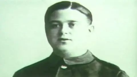 Supplied A black-and-white photograph showing Mooney standing against a plain, light background. He is wearing a dark, formal uniform with a high collar, front buttons, and a wide belt at the waist, with rank-style stripes visible on one sleeve.