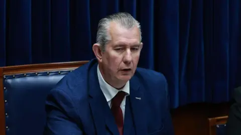 Edwin Poots is wearing a blue suit jacket, red tie and white shirt. He is sitting on the speaker's seat. He has grey, receding hair.