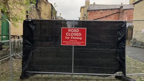 A red sign with white lettering spelling out 'Road Closed for Filming' hanging on a metal fence with a black plastic screen attached to it.