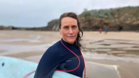 A woman in a blue wetsuit stands on Summerleaze Beach in Bude, Cornwall. She is holding a blue surfboard under her arm. 