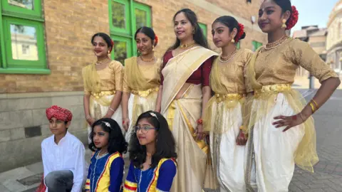 Emma Baugh/BBC A group of women stand in a line wearing matching sarees in gold, white and some red. Two girls wearing a blue, yellow red saree crouch in front of them along with a boy wearing a white shirt and traditional headdress. 