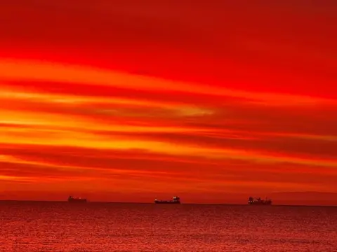 Colette Finlay Three ships float on the water. The entire picture is covered in red light.