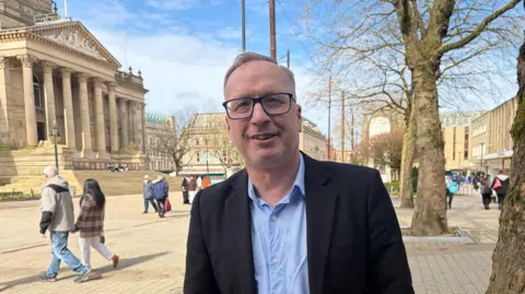 Garry Veevers poses in the public square outside the town hall, which has classically-inspired pillars and front. People walk across the square behind.