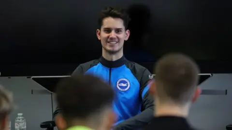 INSTAGRAM The footballer, Toby, is sat and facing a few people, the backs of their heads visible in the image. Toby is wearing blue and back kit for Brighton and is smiling at the people.