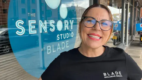 John Fairhall/BBC A head and shoulders image of a woman wearing a black top with the words Blade Inclusive Salon on it. She is looking into the camera.