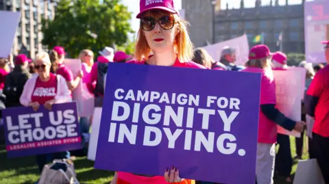 A woman wearing sunglasses in a pink hat holding up a purple placard that says "Campaign for Dignity in Dying". She is at a protest and is surrounded by other women wearing pink hats and clothing.