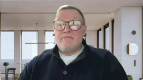 Blockley sitting in a modern, bright room with large windows overlooking the ocean. The space has minimalist décor, including a dining table, contemporary light fixtures, and neutral-toned furniture, giving it an airy, upscale feel.