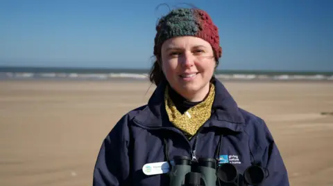 Shaun Whitmore/BBC Hayley Road wearing a multi-coloured hat, yellow spotty neck gaiter and a navy blue jacket. She is stood on a sandy beach below a blue sky. Her brown hair is tied back and she is smiling