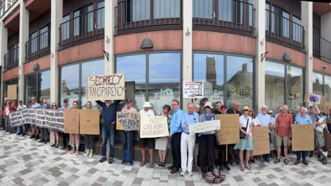 Protestors outside the Cumberland Council building. They are holding various signs saying: "No gas plant dioxins" and the words: "Secrecy Transparency", iwth the words secrecy crossed out.