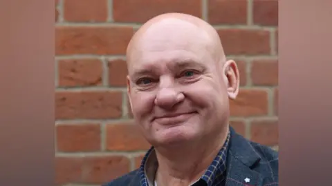 Suffolk Mind A head and shoulders image of a man who is smiling while standing in front of a brick wall.