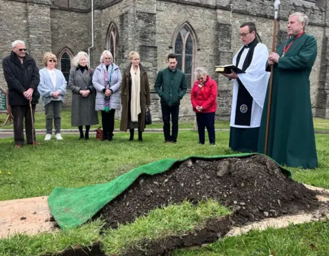 A group of people stand on grass outside a stone church building, gathered around a small mound of earth covered with green fabric. Two people wearing clerical robes stand at the front, one holding an open book, while others stand nearby with heads lowered. 
