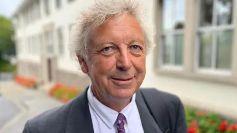 BBC Deputy Bruno Kay-Mouat, a man with whispy grey hair, he's wearing a grey suit and a white shirt, with a purple tie. 