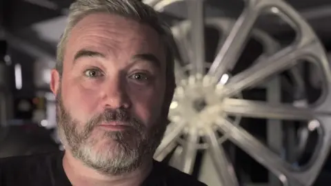 Ewan looks at the camera with a small smile. He has short grey hair and a short grey beard, and wears a black t-shirt. Behind him a projector reel can be seen shining in the dim room. 