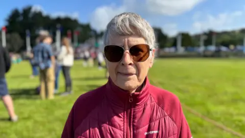 Marian Ashcroft, a woman with short grey hair, she wears sunglasses and a red coat she stands on a field.