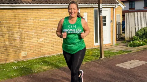 Kim Christon Kim Christon running, with a green top, with her name on it, and black leggings. She is running on a path, with a house behind her. She is smiling with long hair tied back. 