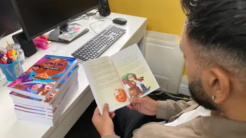 A shot over the shoulder of a seated man who is holding a book open which shows some text and a cartoon drawing of an elderly man sitting in an armchair.