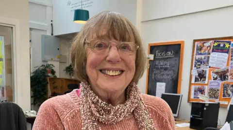 Nina Bale is smiling. She has shoulder length blonde hair a pink jumper and scarf.