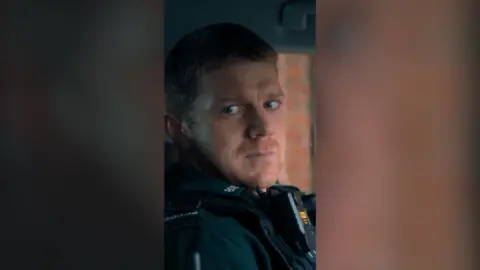 A uniformed police officer in a car, looking wearily to his right. He has short ginger hair and stubble