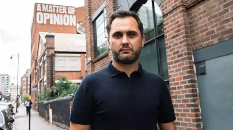 Sam Hufton Man with blue polo shirt and short brown hair standing on a pavement in front of a tall brick building 