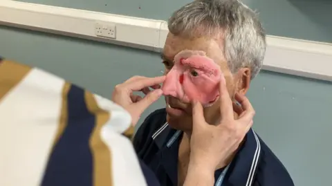 A man with grey hair and wearing a blue polo shirt is seated while a nurse is placing a flesh-like 3D model of half of his face against his left cheek.