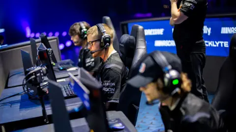 WMGC Esports players take part in a competition. They are sat at desks and laptops sitting in black seats wearing black tops with headphones on.