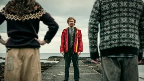 Anthony Dickenson A still from the Incomer shows a man, in a dark grey work suit, green shirt and tie, wearing a bright red anorak, seen through the centre of a man and a woman in knitted jumpers standing with their backs to us.
