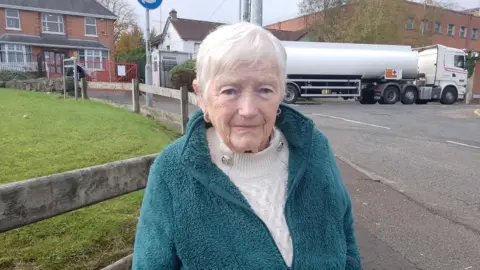 Woman standing on a pavement looking at the camera. Behind her some houses and a oil tanker driving past.