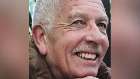 A smiling Geoff King looks up at a point above the camera. He has white hair and a moustache and wears a brown corduroy jacket.