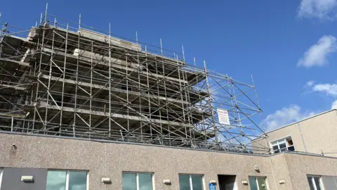 A wide angle view of a grey concrete hospital building. A taller part of it is covered in scaffolding. The larger, nearer section has lots of windows. A blue sky above.