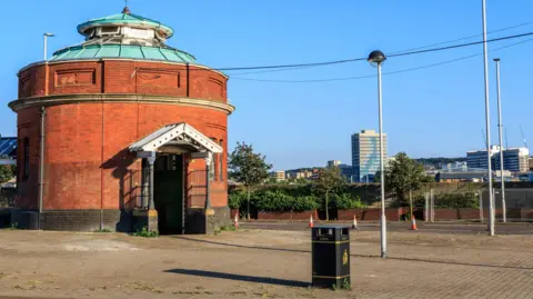 Getty Images The red-brick, domed entrance building to the Woolwich Foot Tunnel stands on an open paved area, with streetlights and modern riverside buildings visible in the background on a clear day.