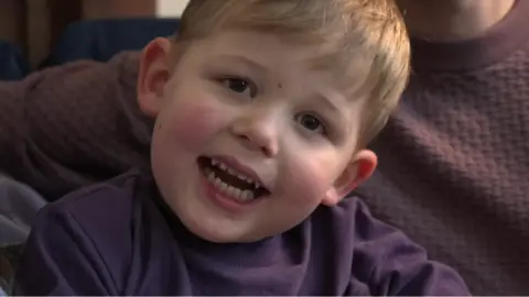 A young boy wearing a purple jumper smiling into the camera