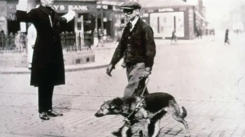 Guide Dogs A black and white image of a man crossing a cobbled road, with a German Shepherd on a lead, a guard in a long black coat, shiny shoes and white gloves stopping traffic. The dog handler is wearing black goggles, trousers with a short jacket, shirt and tie and a flat cap. A Lloyds bank can be seen in the background.