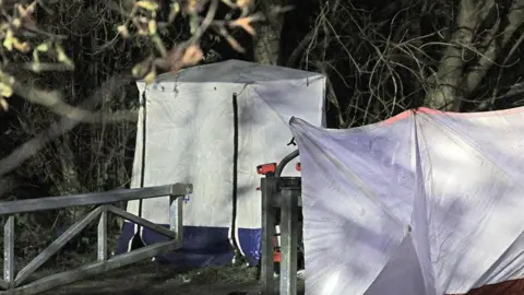 PA Media A white police tent lies near shrubs by a path at the edge of Ashtons Field in the Little Hulton area of Salford. Close by, a metal fence next to a gate is covered with what looks like white plastic sheeting.