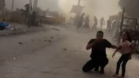 BBC A man and young man stand by a road in the aftermath of an alleged IDF airstrike on a school building.