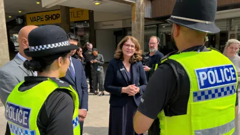 Diana Johnson, policing minister, speaks to two West Midlands Police officers in Coventry city centre. They have their backs to the camera and are wearing yellow high visibility police vests and hard police helmets. There is a female officer to the left and a male officer to the right with the minister in the centre facing towards them.