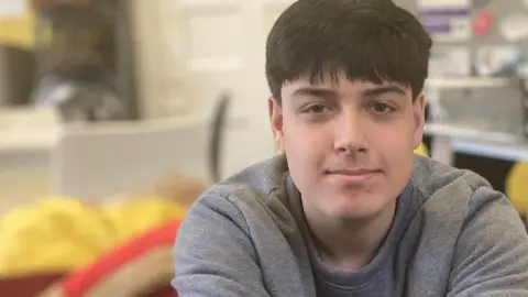 Aaron Smyth, an 18 year old who is sitting on a sofa with red cushions. He is wearing a grey long sleeved T-shirt and is looking directly at the camera and smiling.