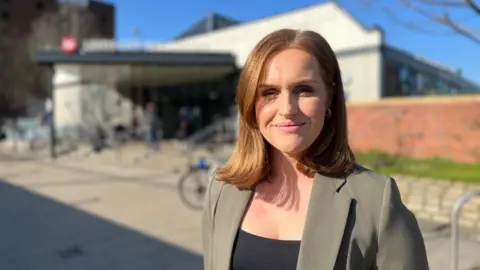 Elizabeth Baines/BBC Woman with shoulder-length auburn hair smiles at the camera. She is pictured wearing a black top and grey/green blazer in front of a bus station which is out of focus.