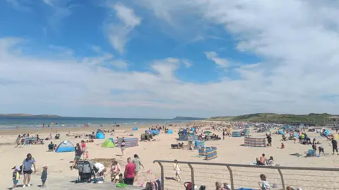 A photo of Portrush beach, with loads of people at the beach and in the sea. Multiple sun shields are up on the sand too. The sun is blue with whispy white clouds and bright sunshine. 