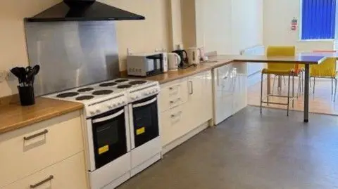 Changing Lives A kitchen with two cookers, three kettles, a microwave and a number of seats and tables.