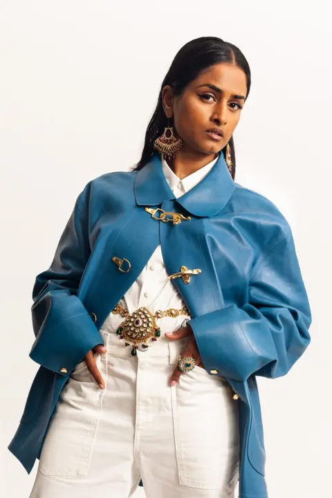 Megan Winston Priya Ragu wore a white dress with a blue overcoat. Her long black hair is tucked behind her ears and she wears bold, gold earrings  