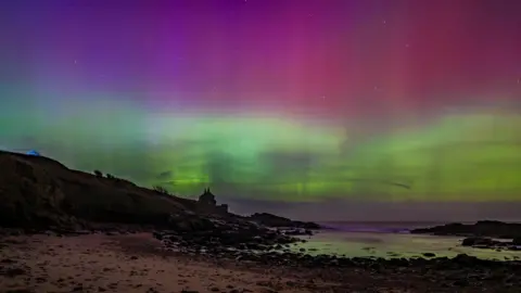Green and pink lights covering the night sky above a beach. The pink looks like it is lying on top of the green in the sky. Stars can be seen underneath the lights. A house can be seen in the distance. The sea is calm with a low tide and stones covering the shoreline.