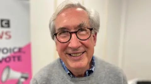 A man with grey hair, brown glasses, wearing a grey jumper. 