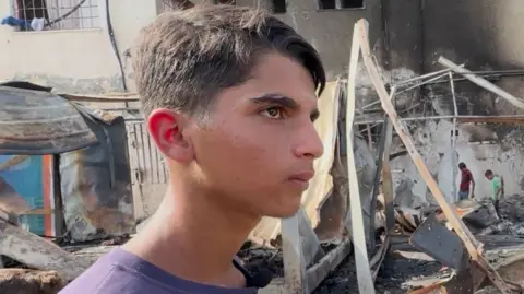 Mohammed al-Dalou, about 17 years old, is a clean-shaven young man wearing a T-shirt. He stands in front of ruined buildings after an airstrike on the al-Aqsa complex in the Gaza Strip. 