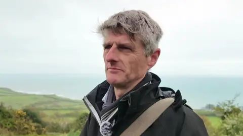 A man with short grey hair is pictured outside. He is stood on coastal cliffs, beyond which we can see the sea and the horizon. He is wearing a black coat and striped scarf and the strap of a bag is slung over his shoulder.