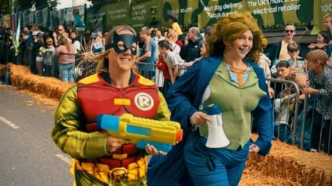 Moussi Productions Two women who are in fancy dress running down a road that is closed as a go-kart race is taking place. Crowds are gathered at the side of the road. The crowd is separated from those involved in the race by metal barriers with hay bales in front of them.