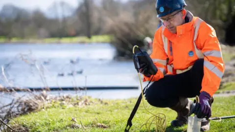 Thames Water A Thames Water worker, wearing an orange hi viz jacket, is performing test by a river. He's wearing a helmet, gloves and is looking at a testing device. It is a sunny day.