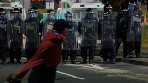 PA Media A man in a red jumper is throwing a brick at a line of armoured police officers