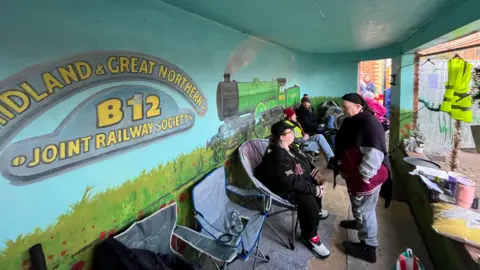 A group of people sit in portable chairs inside a bus shelter with a mural of a steam train on the wall