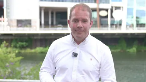 A man in a white shirt is looking directly into the camera. The background is the River Taff and an out of focus shot of the side of the Principality Stadium